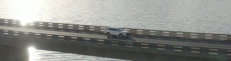 A white car drives over a long, sunlit bridge spanning a vast body of water. The scene is calm and serene, emphasizing the car's solitary journey.